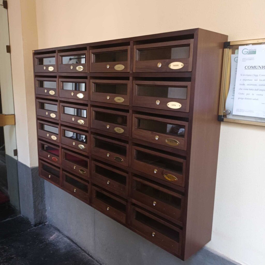 Una bacheca in legno scuro con numerosi caselle postali e targhette dorate all'ingresso di un condominio.