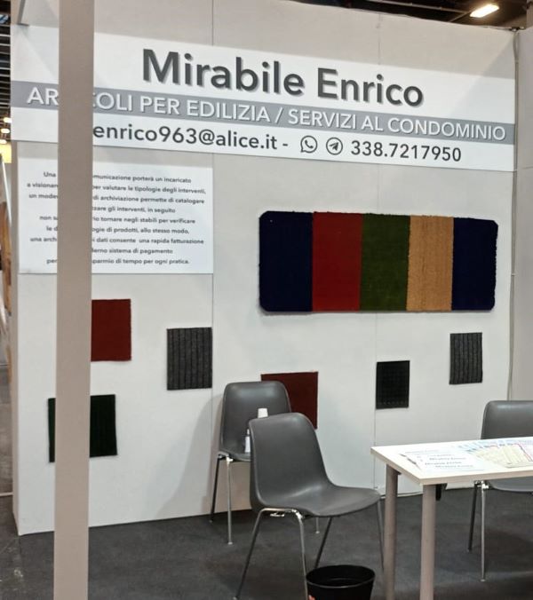 Uno stand fieristico con uno sfondo bianco che riporta il nome "Mirabile Enrico" e il testo "AROLI PER EDILIZIA / SERVIZI AL CONDOMINIO". I dettagli di contatto includono "enrico963@alice.it" e il numero di telefono "338.7217950". Lo stand presenta una varietà di campioni di tessuto o moquette in colori come rosso, verde, beige e blu, montati sulle pareti. Ci sono due sedie grigie, un piccolo tavolo con brochure, una bottiglia di disinfettante per le mani e un cestino nero per i rifiuti sul pavimento.